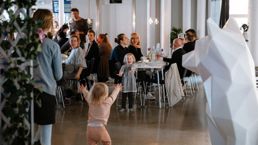 Glada barn springer mot varandra med utsträckta armar i matsalsmiljö. I bakgrunden sitter vuxna vid matbord och samtalar.