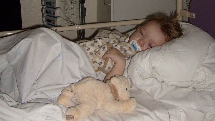 Noel ligger i en sjukhussäng, med napp i munnen, snuttefilt och godsedjur. Foto.