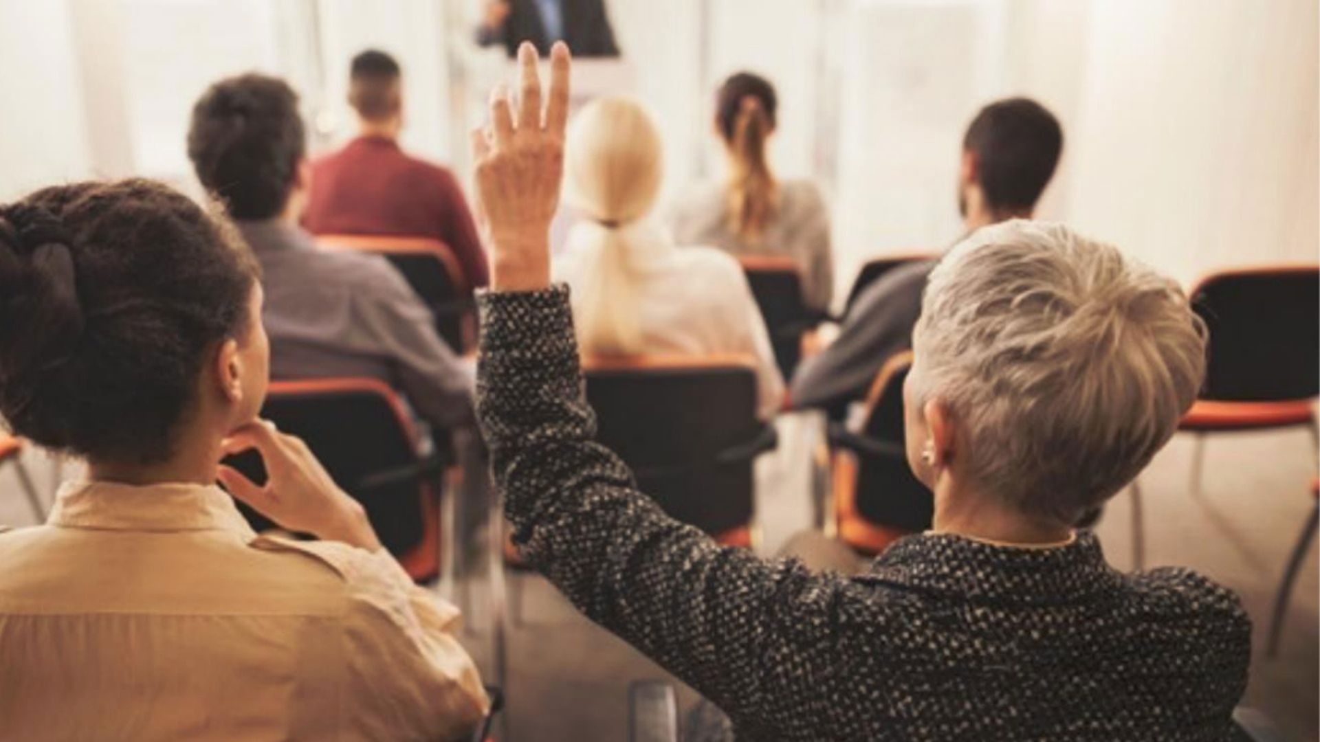Personer bakifrån. Sitter i möte. En person räcker upp handen.
